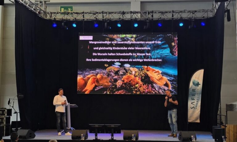Präsentation über die Bedeutung von Mangrovenwäldern auf einer Bühne, zwei Sprecher erläutern ihre Funktion als Sauerstoffproduzenten, Kinderstube für Meerestiere und Wellenbrecher